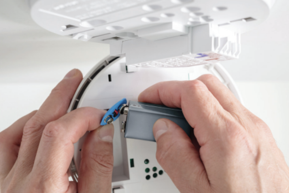 Close-up of hands replacing a rectangular 9-volt battery in a smoke detector mounted on a white ceiling.
